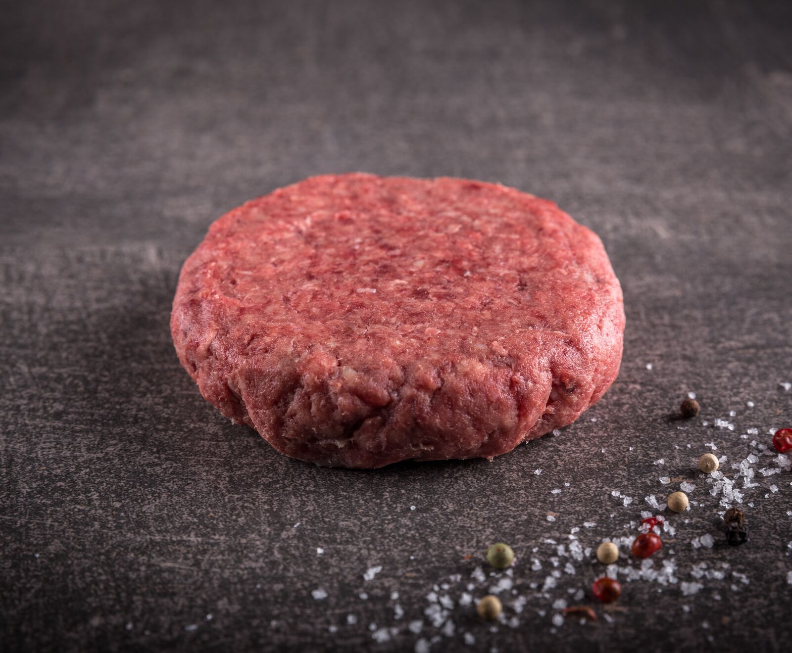 Rohes Rinderhackfleisch-Patty auf dunklem Hintergrund, dekoriert mit Salz und Pfefferkörnern.